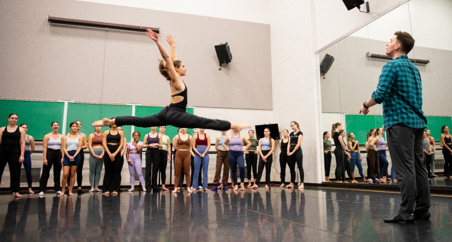 Michael Deeb Weaver offers a dance class, students stand in the background of the studio as a dance leaps across the center of the dance space. 