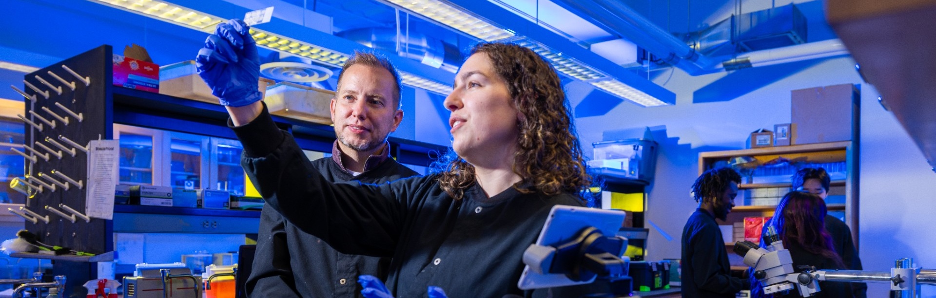 Two researchers wearing lab coats and blue gloves work together in a brightly lit laboratory. One holds up a small sample while explaining it to a colleague, with microscopes, lab equipment, and shelves of supplies visible around them. Other researchers work in the background at lab benches.