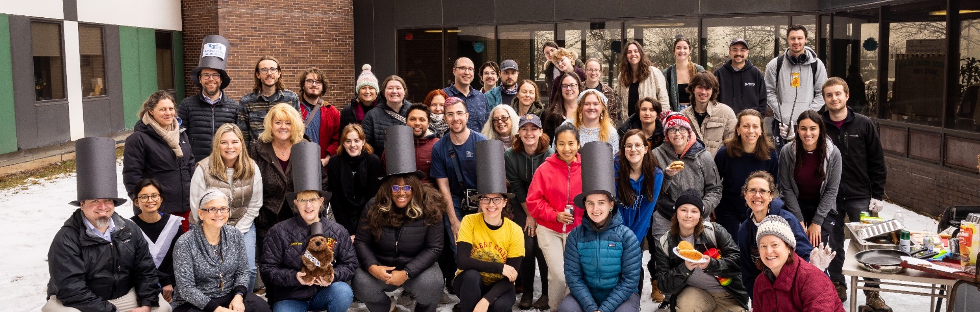 A large group of students, faculty and staff from the Department of Earth Sciences pose outdoors in the snow during the annual Groundhog Day cookout near the Dorsheimer Greenhouse.