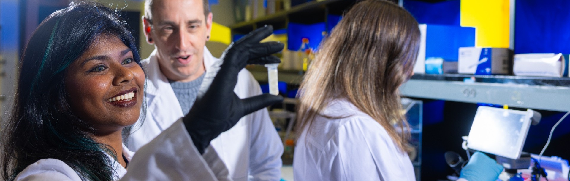 Students work with Gregory Loney in a psychology research lab. One student smiles while holding up a small sample in a vial, wearing gloves and a lab coat, as Loney looks on and gestures. Another student works at a microscope in the background. Lab equipment, shelves, and bright yellow and blue lighting fill the space, illustrating hands-on research and experimentation.