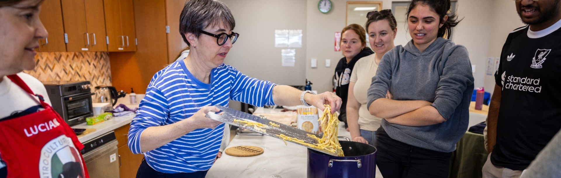 An instructor demonstrates how to cook fresh pasta as students gather around in a teaching kitchen during an Italian language and culture class.