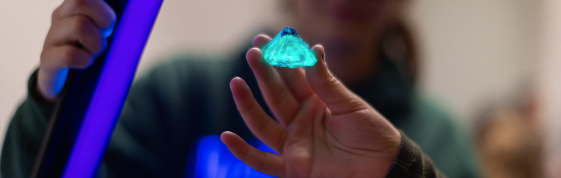 A person holds a small, glowing turquoise crystal under a bright ultraviolet light. The crystal illuminates vividly in the UV beam, while the person holding it is blurred in the background.