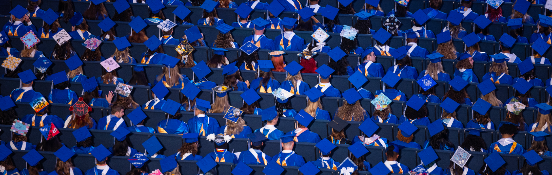 The image is filled with rows of people wearing bright blue graduation caps (mortarboards) and matching gowns. The caps form a repeating geometric pattern across the entire scene. Many caps are plain blue, while others are decorated with colorful designs, messages, or embellishments, adding variation throughout the crowd.